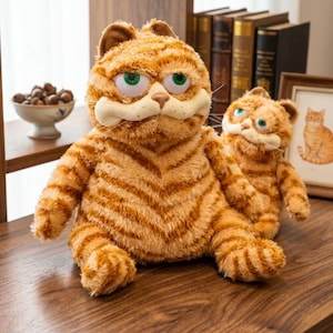May include: Two plush orange tabby cat toys with dark orange stripes, large green eyes, and a light tan muzzle. The larger toy sits in the foreground while a smaller version is positioned behind it, both featuring soft, fuzzy fur textures.