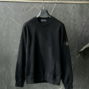 Puede incluir: Una sudadera negra de manga larga con cuello redondo, puños y dobladillo acanalados. Un parche de brújula con bordado amarillo y verde está sujeto a la manga izquierda con dos botones negros. La prenda se muestra colgada en una percha.