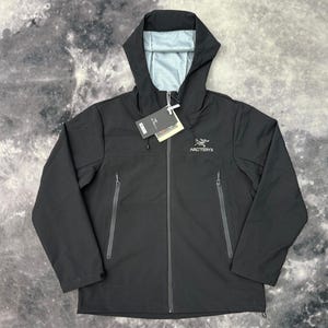 May include: A black hooded jacket featuring a full-length front zip, two vertical zipped hand pockets, and a light grey interior lining. The Arc'teryx logo is embroidered in white on the left chest, and a product tag is attached to the zip.