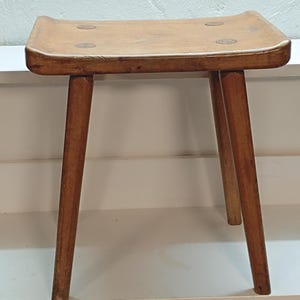 Op de afbeelding: Een kleine houten kruk met een rechthoekige, licht holle zitting voorzien van vier ronde houten inlays op de hoeken. De kruk heeft vier rechte, taps toelopende poten afgewerkt in een middelbruine houttint met een zichtbare houtnerf.