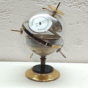 Op de afbeelding: Een bolvormig weerstation met een heldere glazen bol op een ronde messing voet. Binnenin de bol zijn een zilveren thermometerwijzerplaat en een horizontale barometerschaal zichtbaar, samen met dunne messing staven en een kleine schijf.