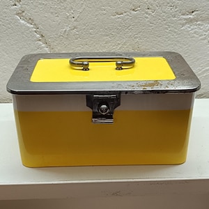 May include: A rectangular metal storage box featuring a bright yellow lid and a yellow body with a white horizontal stripe. The box includes a silver-toned metal handle on top and a central metal latch mechanism on the front for secure closure.