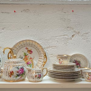 以下が含まれることがあります： 白い磁器をベースに、金色のフィリグリー装飾とカラフルな花柄が施されたティーセット。背の高いティーポット、背の低いティーポット、蓋付きのシュガーボウル、クリーマー、バターディッシュ、複数のソーサー、小さなティーカップが含まれています。