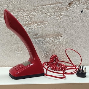 Puede incluir: Un teléfono con cable de color rojo brillante con un auricular curvo y vertical integrado en la base. La base cuenta con una pequeña pantalla digital, varios botones de control cuadrados y un cable rojo en espiral conectado a un enchufe negro.
