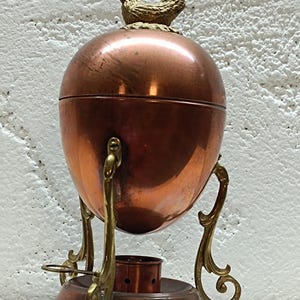 Op de afbeelding: Een koperen eivormige houder met een koperen kippenfiguurtje bovenop, ondersteund door een messing standaard met drie sierlijke poten en een ronde voet. De houder heeft een glad, reflecterend oppervlak en een kleine metalen ring aan één kant.