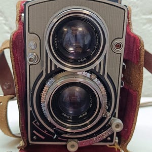 May include: A vintage Flexaret Automat twin-lens reflex camera with a silver metal faceplate, two circular glass lenses, and black dials. The camera is housed in a brown leather case with a deep red velvet interior lining and a leather strap.