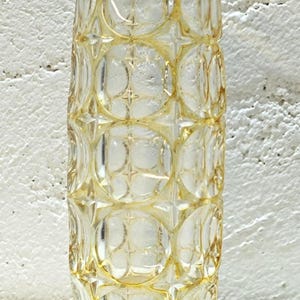May include: A tall, cylindrical glass vase featuring a repeating geometric pattern of raised circles and star-shaped accents. The glass transitions from a clear, transparent body to a vibrant amber-yellow hue at the scalloped rim and base.