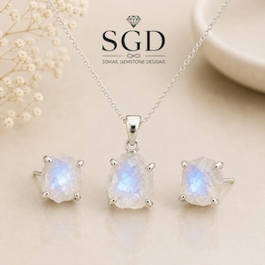 May include: A jewelry set consisting of a silver chain necklace with a faceted, raw-cut moonstone pendant and matching stud earrings. The stones exhibit a blue adularescence. The text SGD SOHAIL GEMSTONE DESIGNS is printed above the pendant.