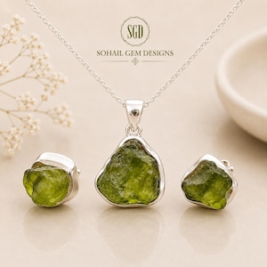 Può includere: Un set di gioielli composto da una collana a catena in argento con un ciondolo in pietra grezza verde di forma irregolare e orecchini a lobo abbinati. Il testo SOHAIL GEM DESIGNS è centrato sopra. Le pietre hanno una superficie ruvida.