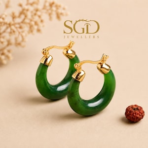 Peut inclure: Une paire de boucles d'oreilles créoles composées d'anneaux en jade vert foncé poli fixés à des fermoirs en métal doré. Les éléments métalliques portent le poinçon 585. Le texte SGD JEWELLERS est visible au-dessus dans une police stylisée.