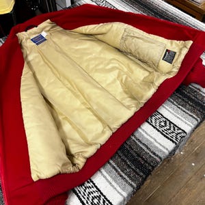 May include: A red bomber jacket with a beige quilted lining. The jacket has a ribbed collar, cuffs, and hem. The interior features a brand tag. The jacket is open, revealing the interior.