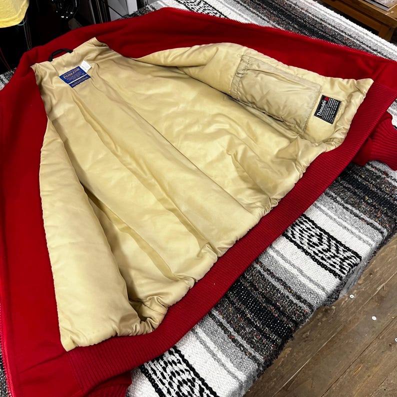 May include: A red bomber jacket with a beige quilted lining. The jacket has a ribbed collar, cuffs, and hem. The interior features a brand tag. The jacket is open, revealing the interior.