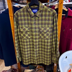 May include: A green and brown plaid shirt with a button-down front and two chest pockets. The shirt has a collar and the brand name "Pendleton" is visible on a tag. The shirt is displayed on a wooden rack, with other clothing items visible.