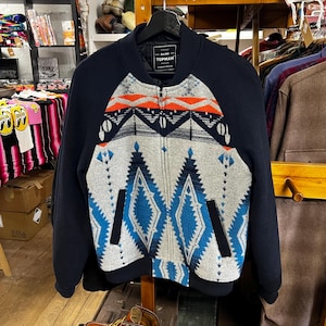 May include: A navy blue bomber jacket with a geometric pattern in gray, orange, and blue. The jacket has a zip-up front, side pockets, and the brand name "TOPMAN" on the label.