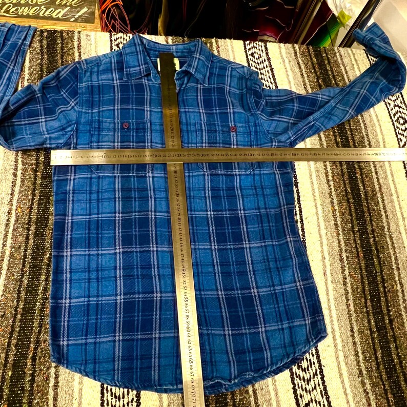 May include: A blue plaid button-down shirt with long sleeves. The shirt features a classic collar, two chest pockets, and red buttons. A ruler is placed across the shirt to measure its length. The shirt is made of a soft, woven fabric.