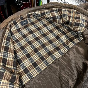 May include: Brown leather jacket with a plaid interior in shades of brown, black, and cream. The jacket has a brown satin lining and a tag that reads "saki."
