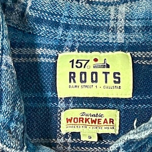 May include: Close-up of a blue and white plaid shirt with two tags. The top tag reads "157 ROOTS Dairy Street 1 G'allstad". The bottom tag says "Durable WORKWEAR MADE TO FIT - FIT TO WEAR".
