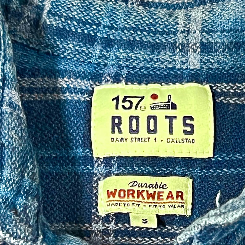 May include: Close-up of a blue and white plaid shirt with two tags. The top tag reads "157 ROOTS Dairy Street 1 G'allstad". The bottom tag says "Durable WORKWEAR MADE TO FIT - FIT TO WEAR".
