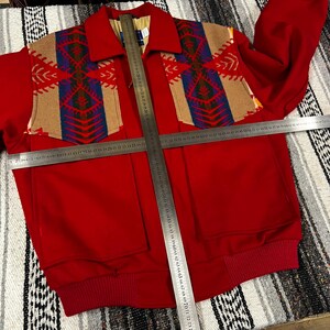 May include: A red jacket with a patterned design on the shoulders and upper chest. The design features tan, blue, and red geometric shapes. The jacket has two front pockets and a ribbed hem. The jacket's length is approximately 71 cm.