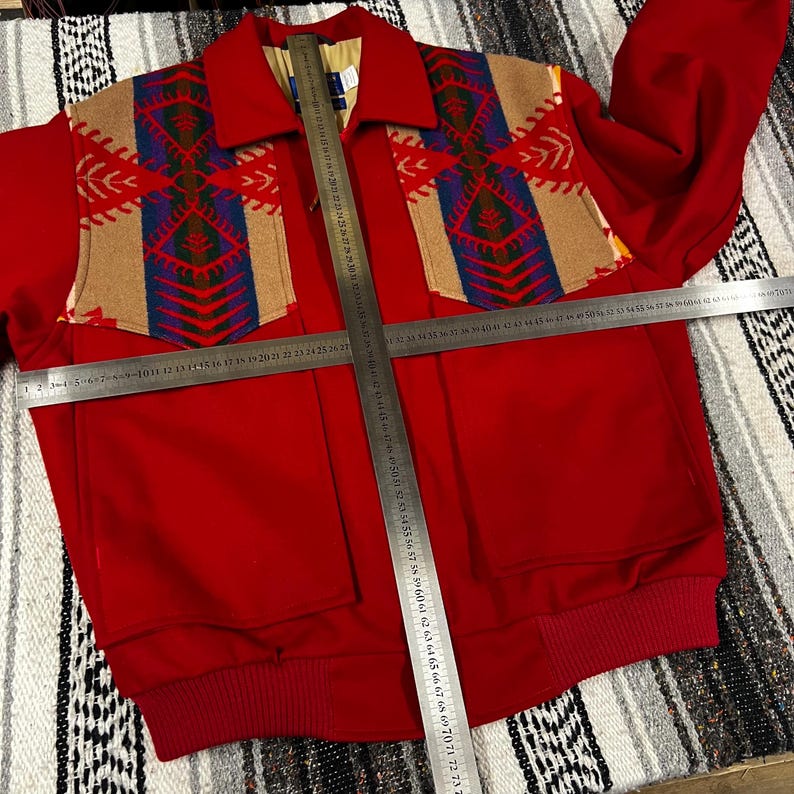 May include: A red jacket with a patterned design on the shoulders and upper chest. The design features tan, blue, and red geometric shapes. The jacket has two front pockets and a ribbed hem. The jacket's length is approximately 71 cm.