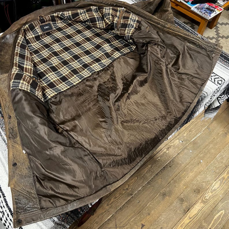 May include: Open brown leather jacket with a plaid lining in shades of brown, black, and cream. The jacket is unbuttoned, revealing the interior lining and the texture of the leather.