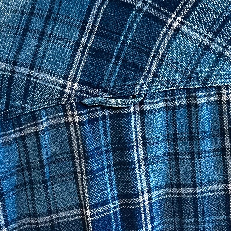 May include: Close-up of a blue plaid fabric. The pattern features a grid of intersecting lines in shades of blue and white. The fabric appears to be a woven material, possibly cotton or a similar textile. The image highlights the texture and color variations.