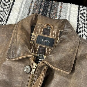 May include: Close-up of a brown leather jacket with a collar and a zipper. The jacket has a worn, distressed appearance. The interior lining is patterned with brown and beige herringbone fabric. The brand name "saki." is visible on a tag.