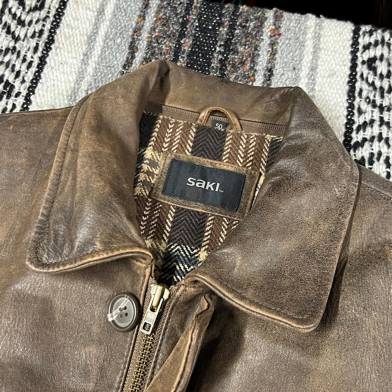 May include: Close-up of a brown leather jacket with a collar and a zipper. The jacket has a worn, distressed appearance. The interior lining is patterned with brown and beige herringbone fabric. The brand name "saki." is visible on a tag.