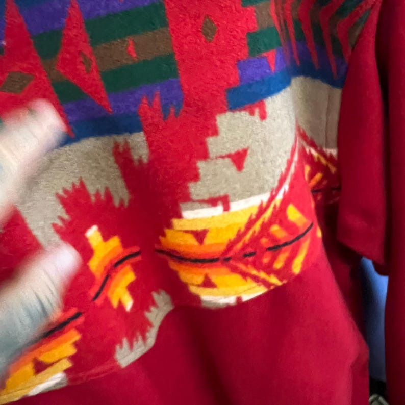 May include: A red sweater with a colourful geometric pattern across the shoulders and upper chest. The pattern includes shades of red, brown, purple, green, and yellow. The sweater has long sleeves.