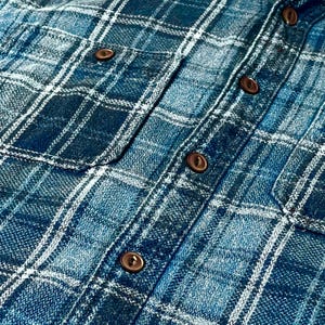 May include: Close-up of a blue plaid shirt with brown buttons. The shirt has two chest pockets and a classic button-down design. The plaid pattern features shades of blue and white.