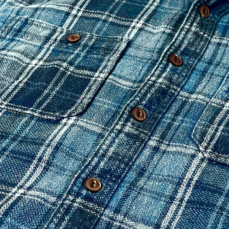 May include: Close-up of a blue plaid shirt with brown buttons. The shirt has two chest pockets and a classic button-down design. The plaid pattern features shades of blue and white.