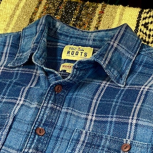 May include: Close-up of a blue plaid shirt with a button-down collar. The shirt features a classic plaid pattern in shades of blue and white. The label reads "157 ROOTS" and "WORK".