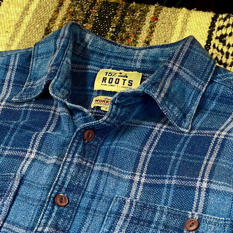 May include: Close-up of a blue plaid shirt with a button-down collar. The shirt features a classic plaid pattern in shades of blue and white. The label reads "157 ROOTS" and "WORK".