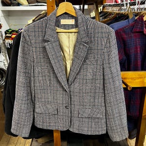May include: A gray and black plaid blazer with a notched collar, two front pockets, and a single-button closure. The blazer is hanging on a wooden hanger. The interior lining is a light tan color.