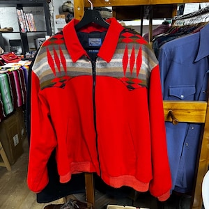 May include: A red zip-up jacket with a patterned yoke in shades of gray, brown, and green. The jacket has a collar and ribbed cuffs and hem. The jacket is hanging on a black hanger.