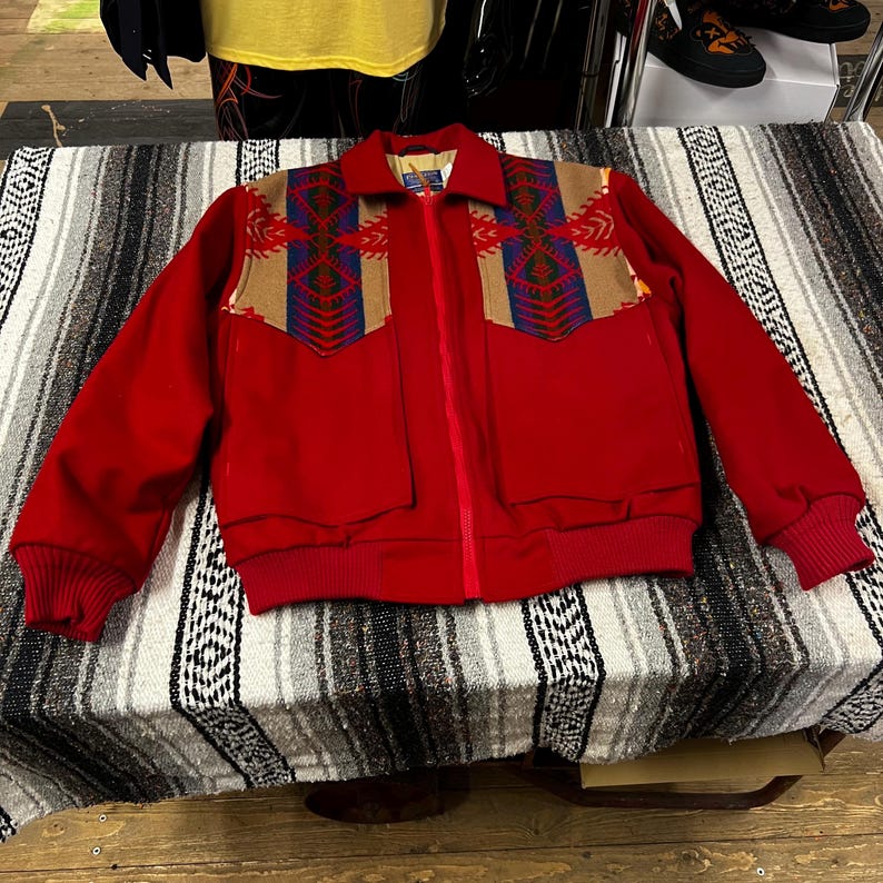 May include: A bright red jacket with a full-length zip and two front pockets. The jacket has tan and patterned shoulder panels with blue, red, and green geometric designs. The cuffs and waistband are ribbed.