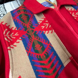 May include: A red wool jacket with a full zip and a tan and patterned front panel. The panel features a geometric design in red, blue, purple, and green. The jacket has a collar and a tag that says "PENDLE".
