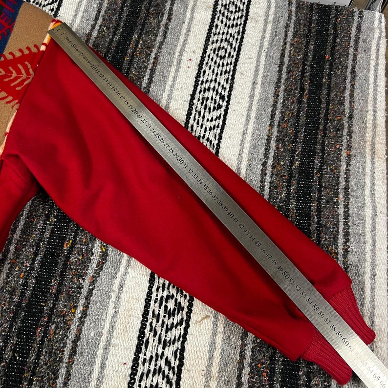 May include: A red long-sleeved sweater with a ribbed cuff. A metal ruler is placed across the sweater, measuring approximately 152 cm. The sweater is on a patterned blanket.