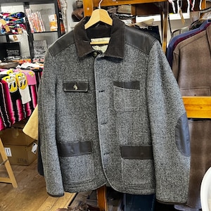 May include: A grey tweed jacket with leather detailing on the shoulders, elbows, and pockets. It features a brown corduroy collar and a button-up front. The jacket is suspended from a wooden hanger.
