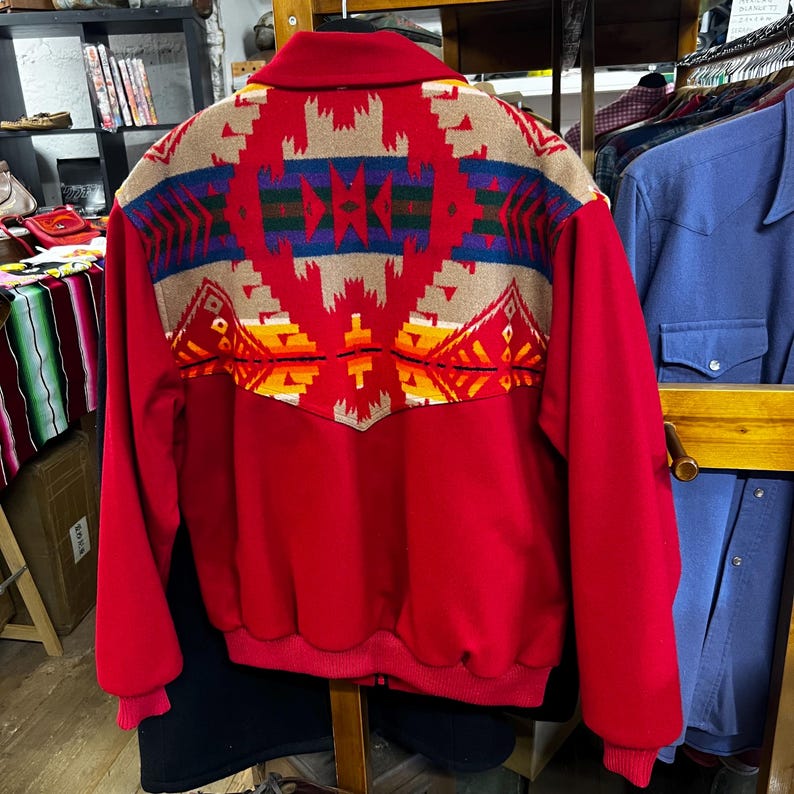 May include: A red bomber jacket with a colourful geometric design on the back. The design features shades of tan, blue, green, yellow, and orange. The jacket has a zip-up front and a collar.