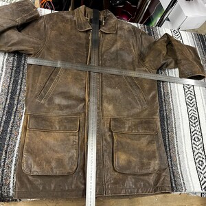 May include: A brown leather jacket with two large front pockets and a collared neckline. The jacket has a distressed appearance and a full-length zipper. A metal ruler is placed across the jacket. The jacket appears to be a medium size.