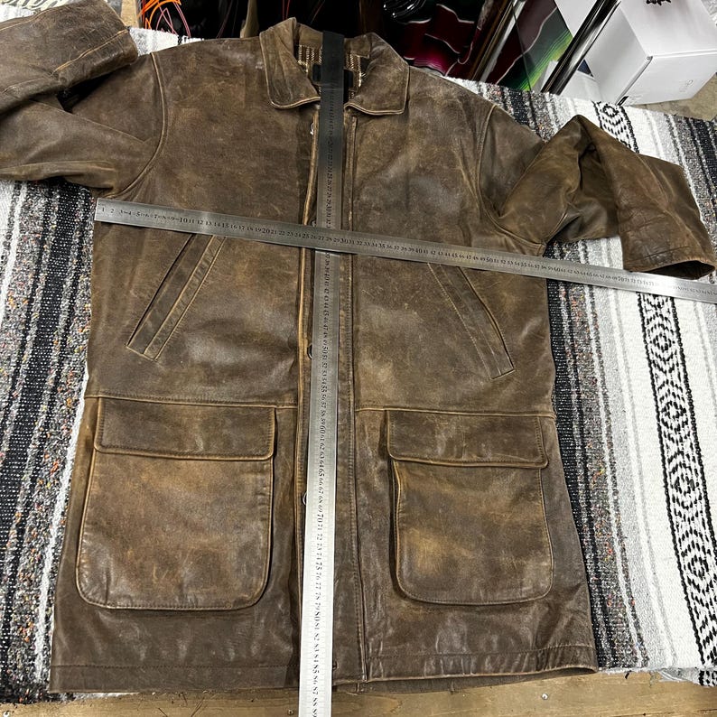 May include: A brown leather jacket with two large front pockets and a collared neckline. The jacket has a distressed appearance and a full-length zipper. A metal ruler is placed across the jacket. The jacket appears to be a medium size.