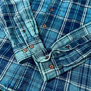 May include: Close-up of a blue plaid flannel shirt. The shirt features a classic button-down design with brown buttons and cuffs. The fabric has a textured appearance, with a mix of light and dark blue hues.