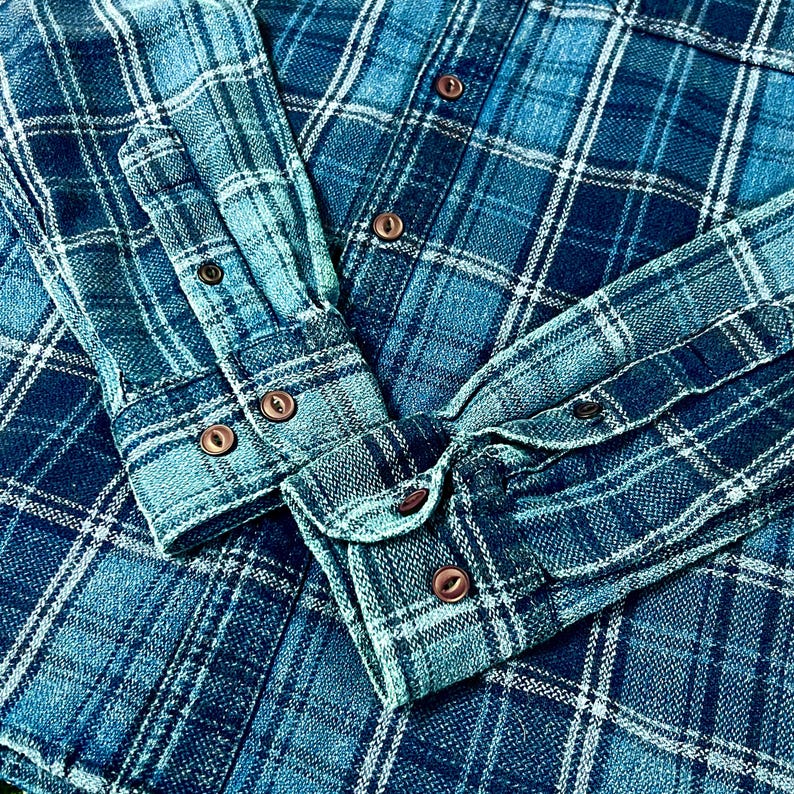 May include: Close-up of a blue plaid flannel shirt. The shirt features a classic button-down design with brown buttons and cuffs. The fabric has a textured appearance, with a mix of light and dark blue hues.