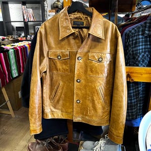 May include: A vintage, tan leather jacket with a classic collar and button-up front. The jacket features two chest pockets and two side pockets. The jacket is hanging on a black hanger. The brand name "CONWELL" is visible on the label.