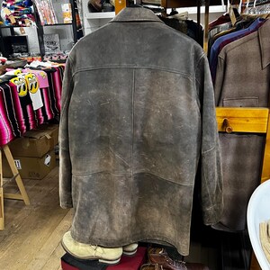 May include: A vintage brown leather jacket with a classic collar and button closure. The jacket has a worn, distressed appearance, with visible texture and variations in color. The back of the jacket is visible, showcasing the seams and design details.
