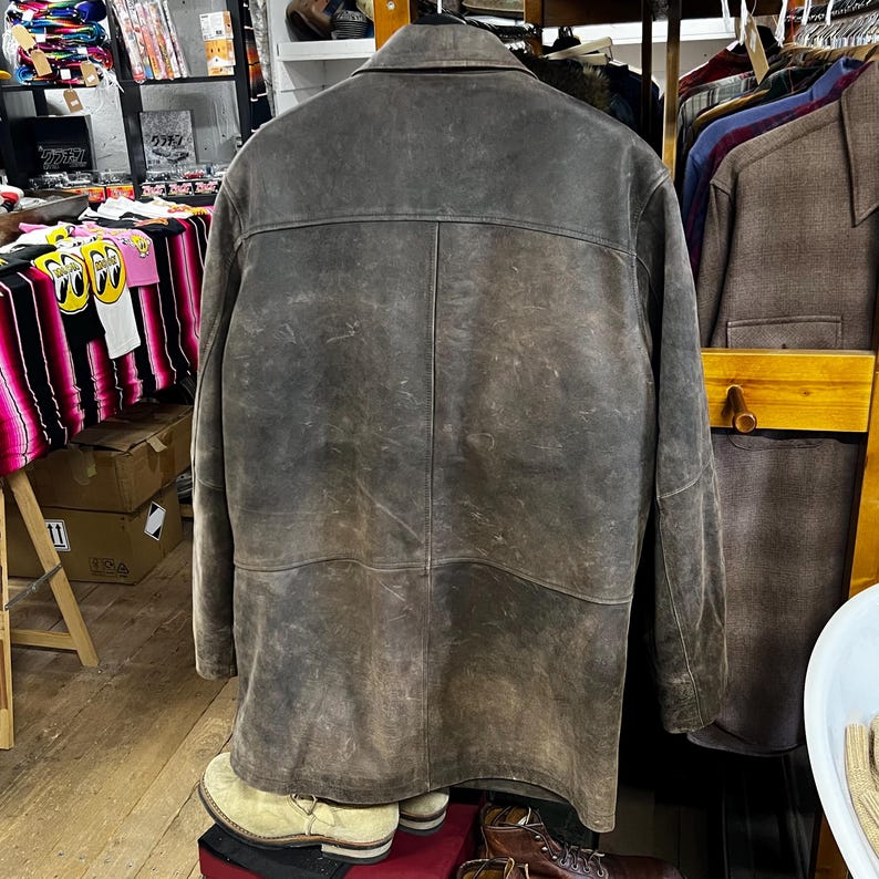 May include: A vintage brown leather jacket with a classic collar and button closure. The jacket has a worn, distressed appearance, with visible texture and variations in color. The back of the jacket is visible, showcasing the seams and design details.