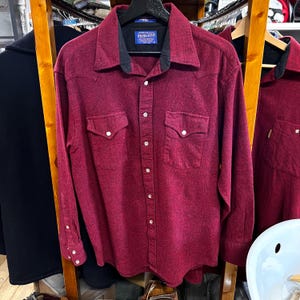 Puede incluir: Una camisa burdeos de manga larga con cuello negro y dos bolsillos en el pecho. La camisa tiene botones blancos y el nombre de la marca "Pendleton" en una etiqueta. La tela es texturizada.