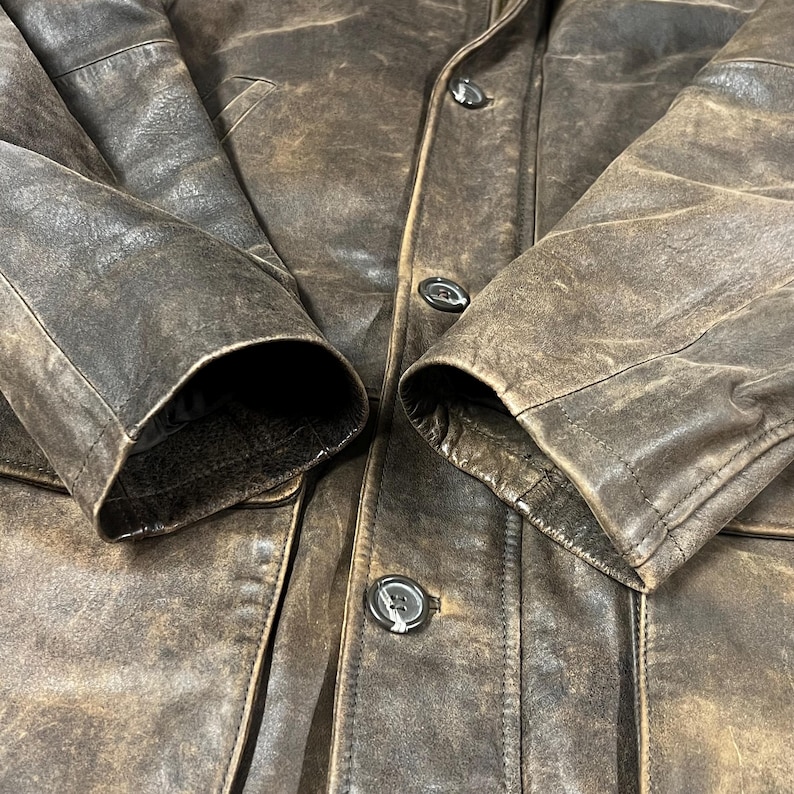 May include: Close-up of a brown leather jacket with button closures. The jacket has a distressed, weathered appearance, with visible texture and variations in color. The sleeves are visible, showing the cuffs and stitching details.