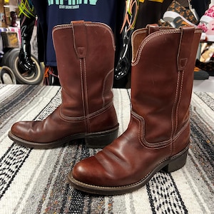 Könnte beinhalten: Ein Paar braune Leder-Cowboy-Stiefel mit weißen Nähten. Die Stiefel haben ein klassisches Western-Design mit spitzer Zehenpartie, einem mittelhohen Schaft und einem gestapelten Absatz. Das Leder hat ein abgenutztes Aussehen.